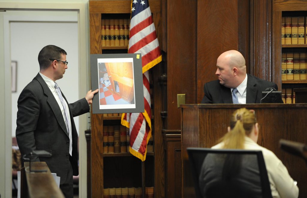 From left, defense attorney Patrick Noonan, shows East Bridgewater police officer Peter Belmore, a photograph of the couples home a door panel leading into the laundry room that seemed to be kicked out. The trial for a former Boston firefighter accused of assaulting his wife began Tuesday in Brockton Superior Court.<br /> Richard R. Parker, who worked for the Boston Fire Department for nearly 26 years and was also a 9/11 responder, faces a charge of assault to murder amid others against his wife, Kimberly Boleza Parker, from an alleged incident in 2011. Parker died March 10, 2013 of causes that have not yet been determined.<br /> On Dec. 18, 2011, at 5 a.m., police responded to a 911 call placed from their Satucket Avenue home reporting that Parker was “threatening his wife with a knife,” police logs show.<br /> In a written testimony following the incident, Kimberly Parker described a violent scene in which Parker was throwing knives at her while she stood pinned against a bedroom wall, according to court records.<br /> She was able to escape, she wrote, after one knife took a bad bounce and landed out of sight.<br /> Parker, 58, of 3 Satucket Ave., was indicted in 2012 following his wife’s testimony in front of a grand jury. He is charged with kidnapping; assault to murder; assault with a dangerous weapon, a knife; threat to commit a crime and intimidation of a witness.<br /> Police, on two prior occasions before 2011, responded to the home for abuse reports.<br /> Kimberly Parker died March 10, 2013 at age 45 in her East Bridgewater home of causes that have not yet been determined.<br /> (Marc Vasconcellos/The Enterprise)