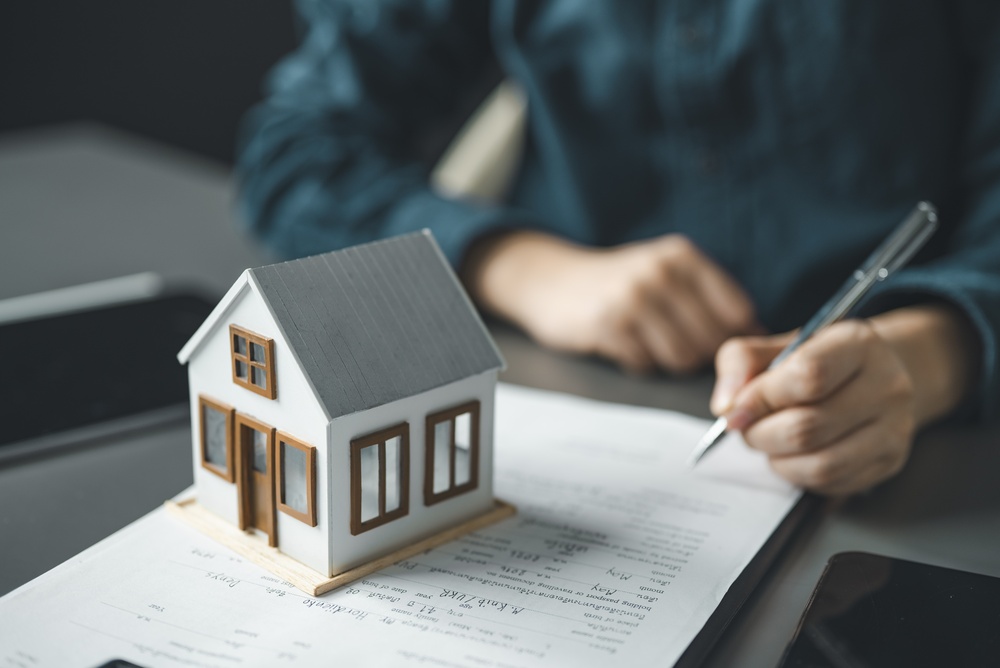 Small model house sitting on top of property documents while a person signs paperwork